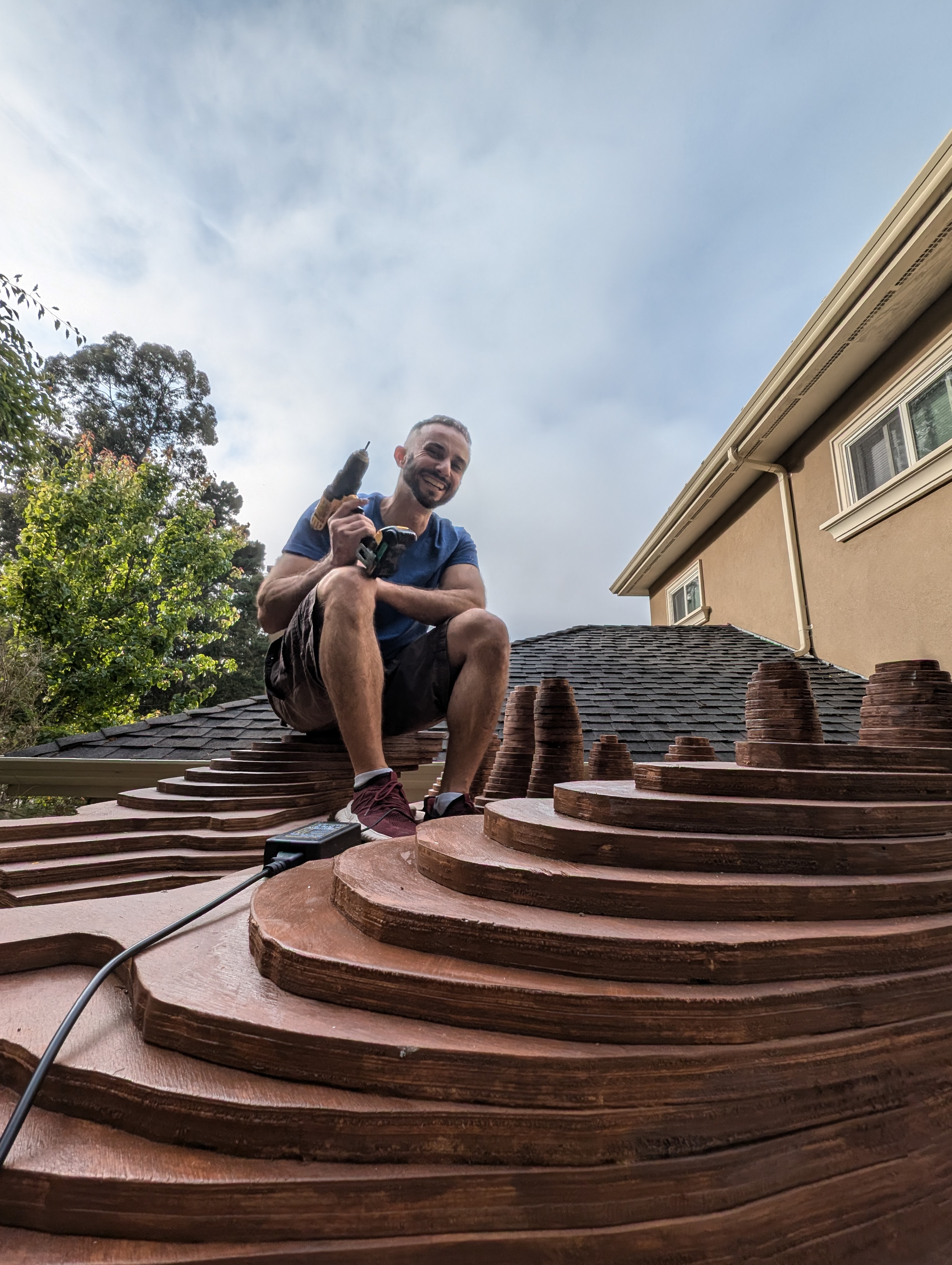 The artist working on a wooden sculpture, sitting on layered wooden elements with a power tool